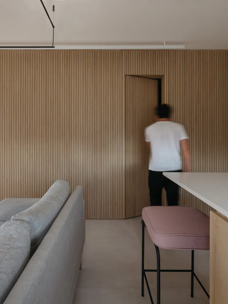 A modern residential kitchen featuring wood-paneled walls, a contemporary sofa, and a minimalist barstool with pink cushions.