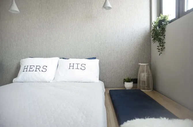 A minimalist bedroom featuring a queen-sized bed with 'Hers' and 'His' pillows, a blue cushion, a window with greenery, and a