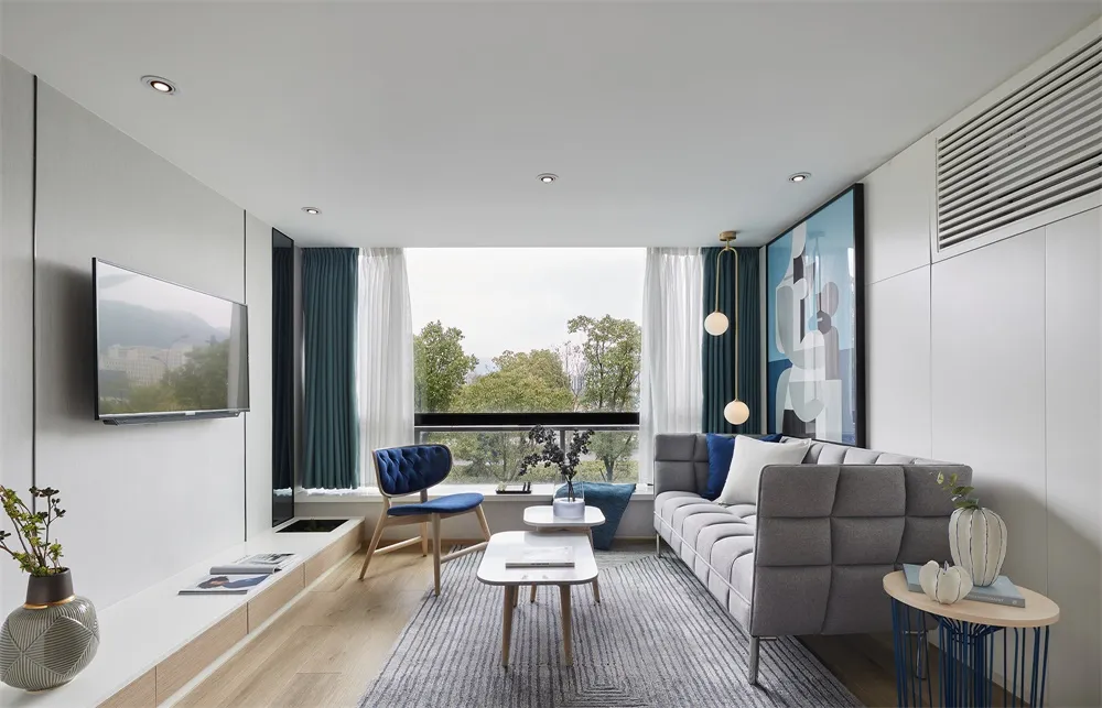 Modern, minimalist living room featuring a large window with greenery outside, a grey sofa, and blue accents.