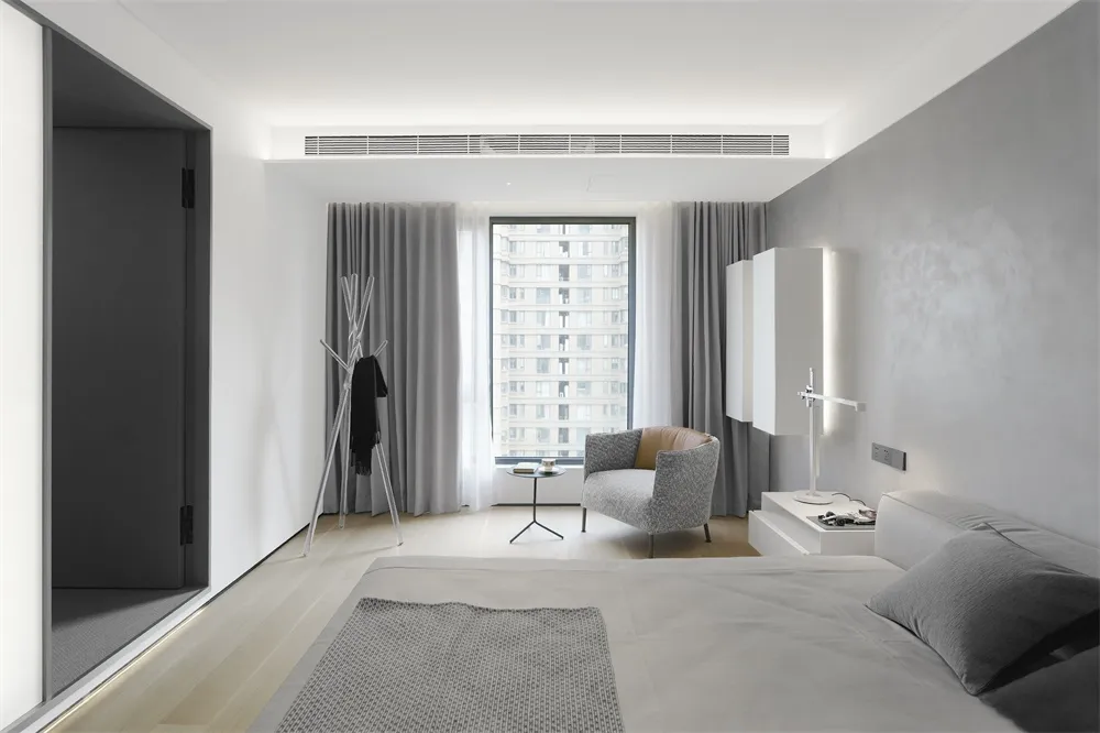 A modern bedroom with a minimalist design, featuring a bed, armchair, and window with curtains, all in neutral tones.