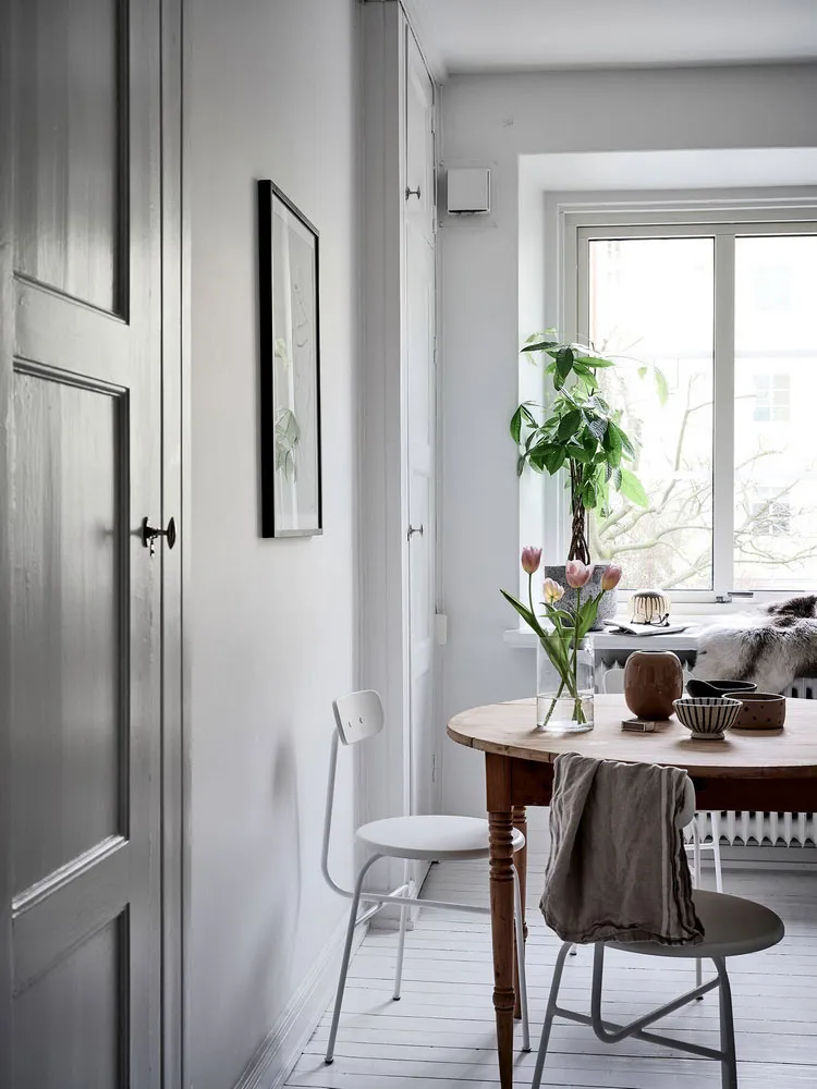 Modern, white minimalist interior with wood dining table and chairs, minimalist art, and a plant on the window sill.