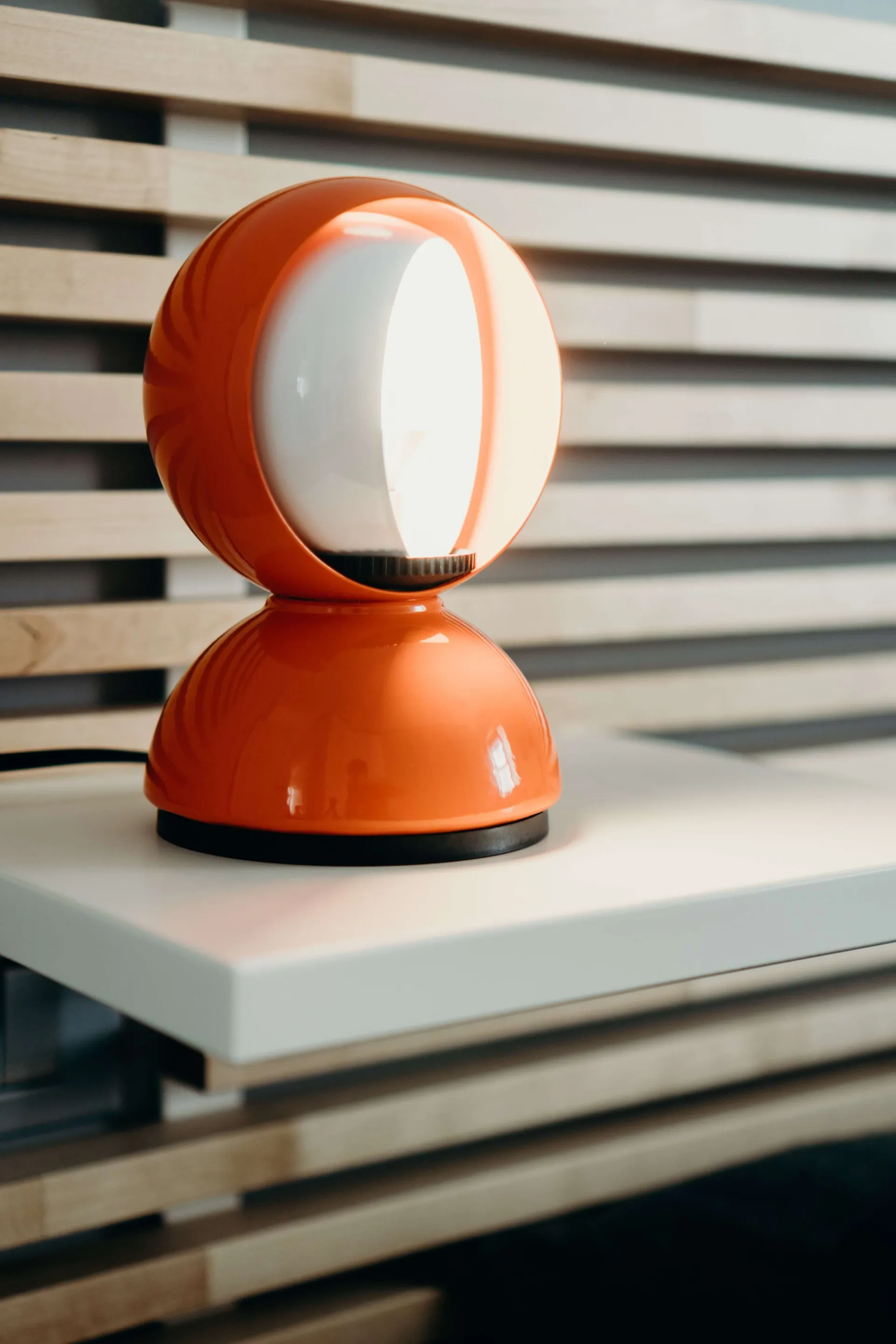 An orange table lamp with a white light bulb inside, placed on a white surface with light wood slats in the background.