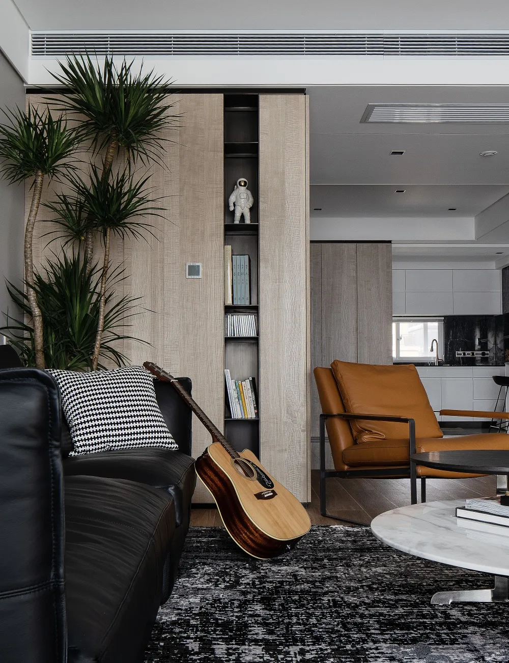 A modern living room with a wooden accent wall, a black leather couch, a brown leather armchair, and a guitar leaning against