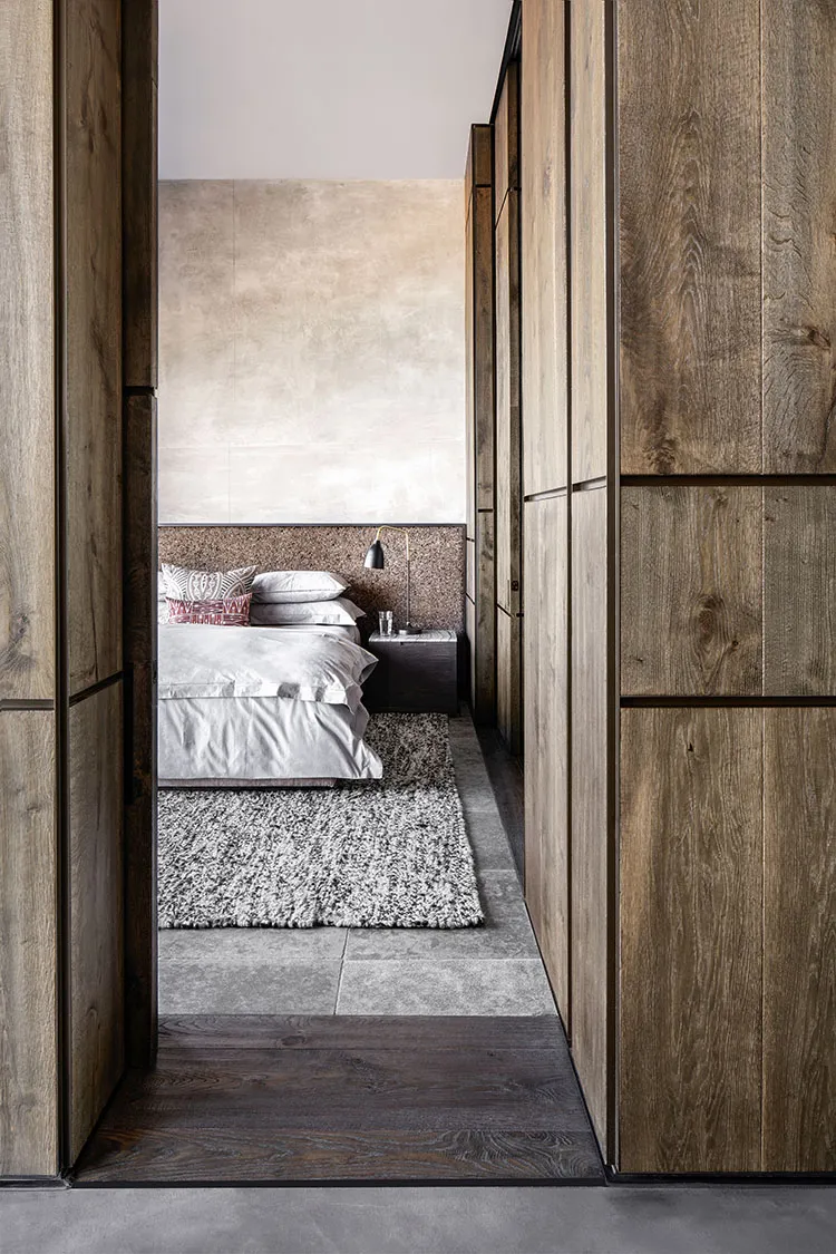 Modern minimalist bedroom with wood-paneled walls, concrete tiles, a bed with a soft bedspread and patterned pillows, and a b