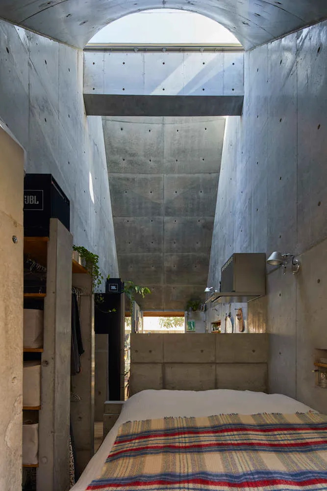 A minimalist bedroom featuring a concrete wall and floor, with a bed covered in a striped blanket and a skylight above.