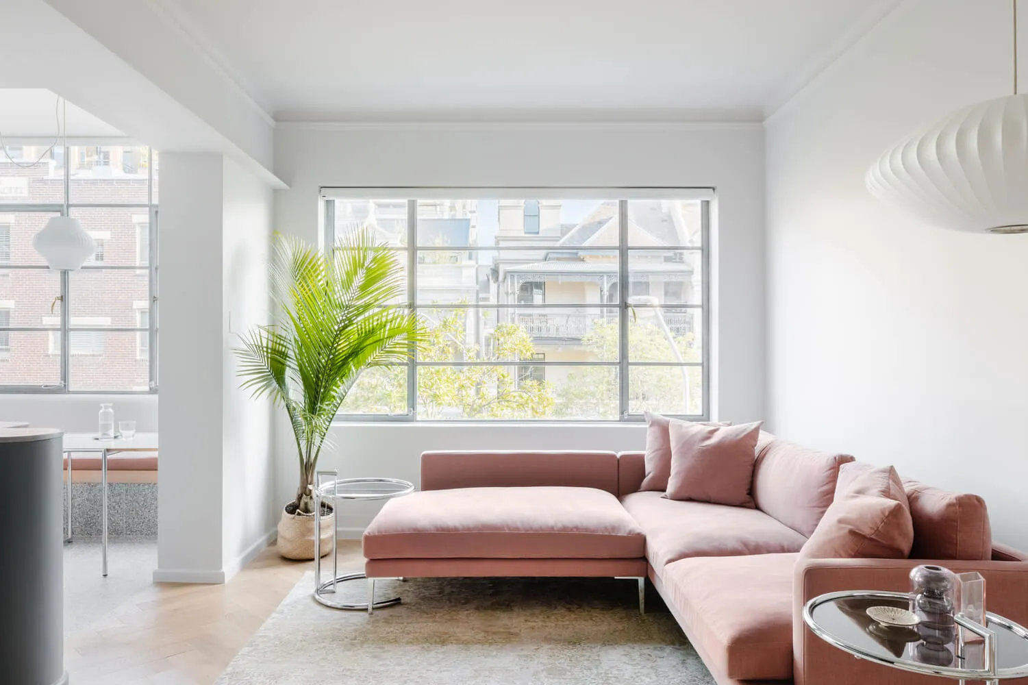 A modern, minimalist living room with a pink sofa, glass table, and large window overlooking urban buildings.