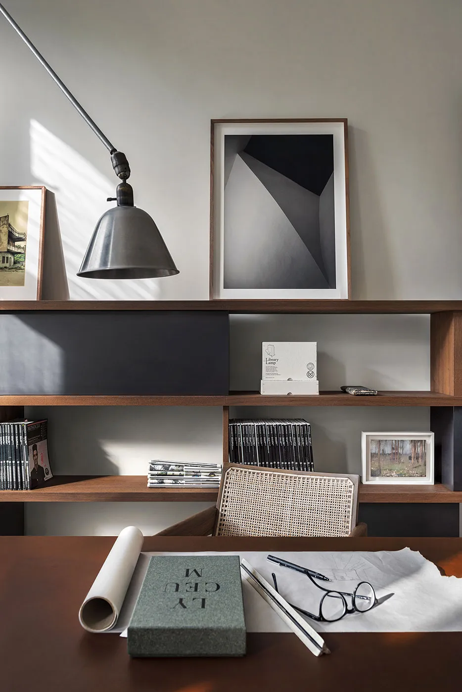 A modern office workspace featuring a wooden shelving unit with books, a sleek metal lamp, and various framed artworks.
