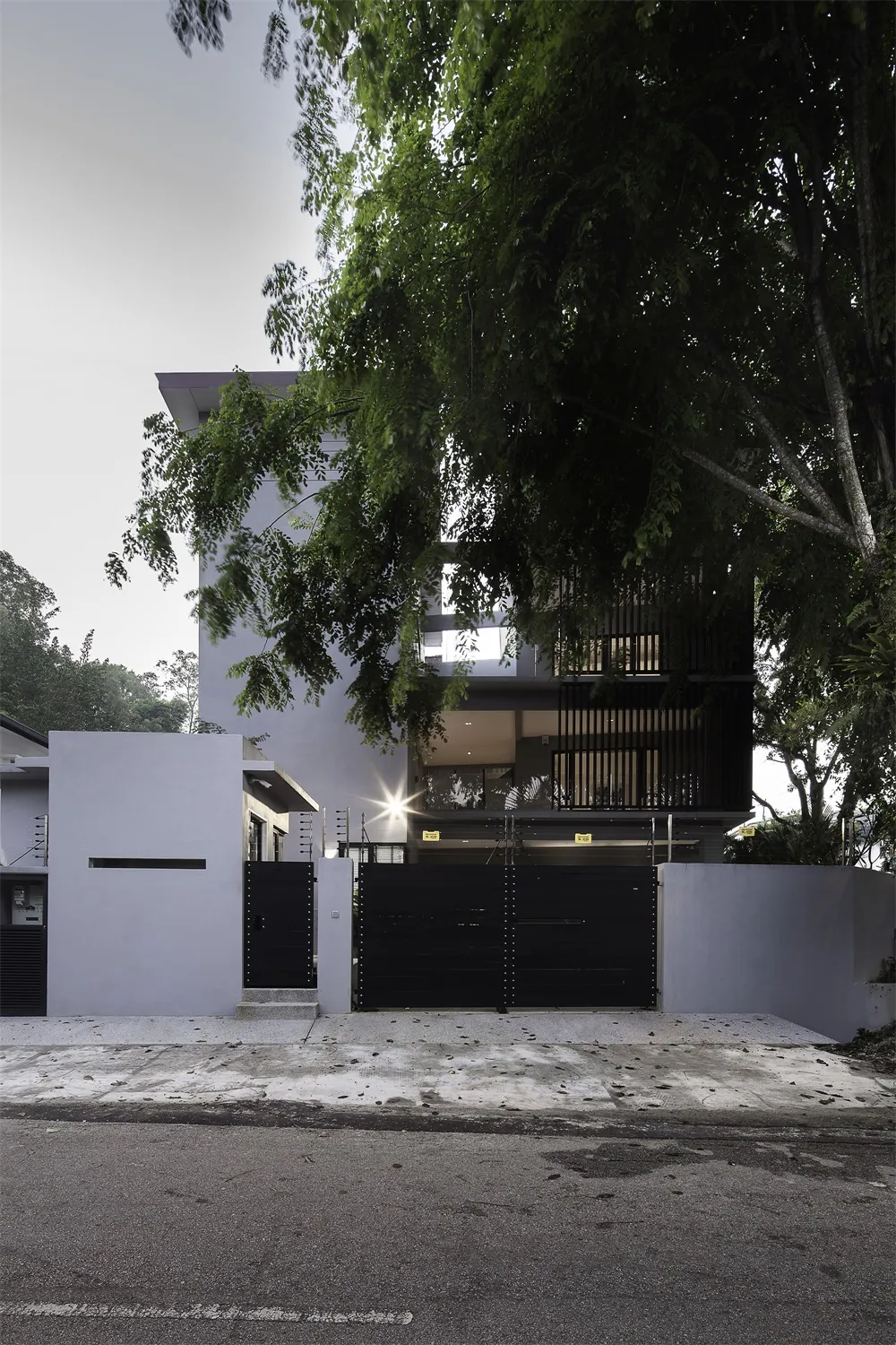 A modern residential building with a concrete and glass exterior, featuring a secure black gate and surrounded by lush greene