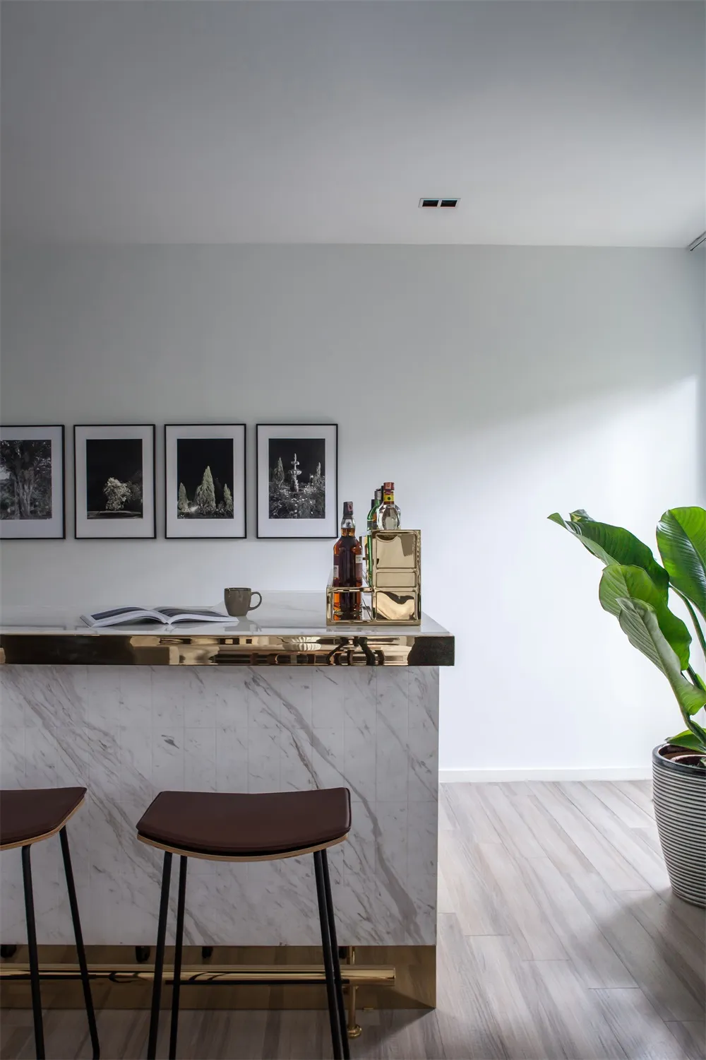 A minimalist bar studio featuring a marble counter with stools and a shelf of bottles and books.
