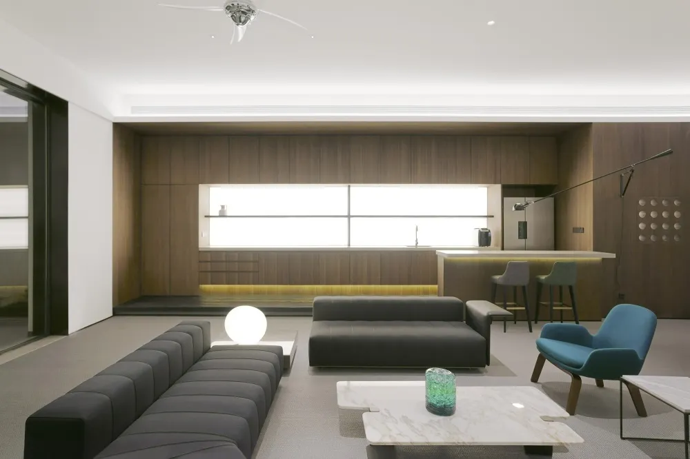 Modern living room with dark sofas, marble coffee tables, wood-paneled walls, and glass bar setup