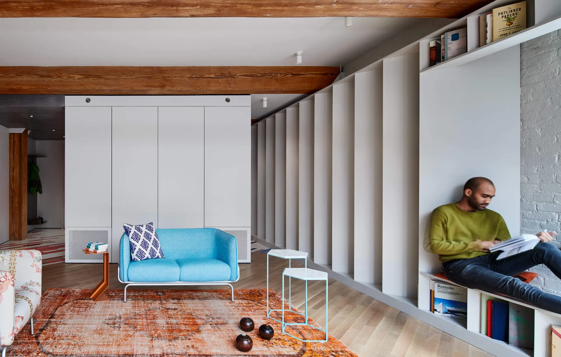 A minimalist living room with a blue sofa, side table, and rug, featuring a bookshelf with books and a man sitting and readin