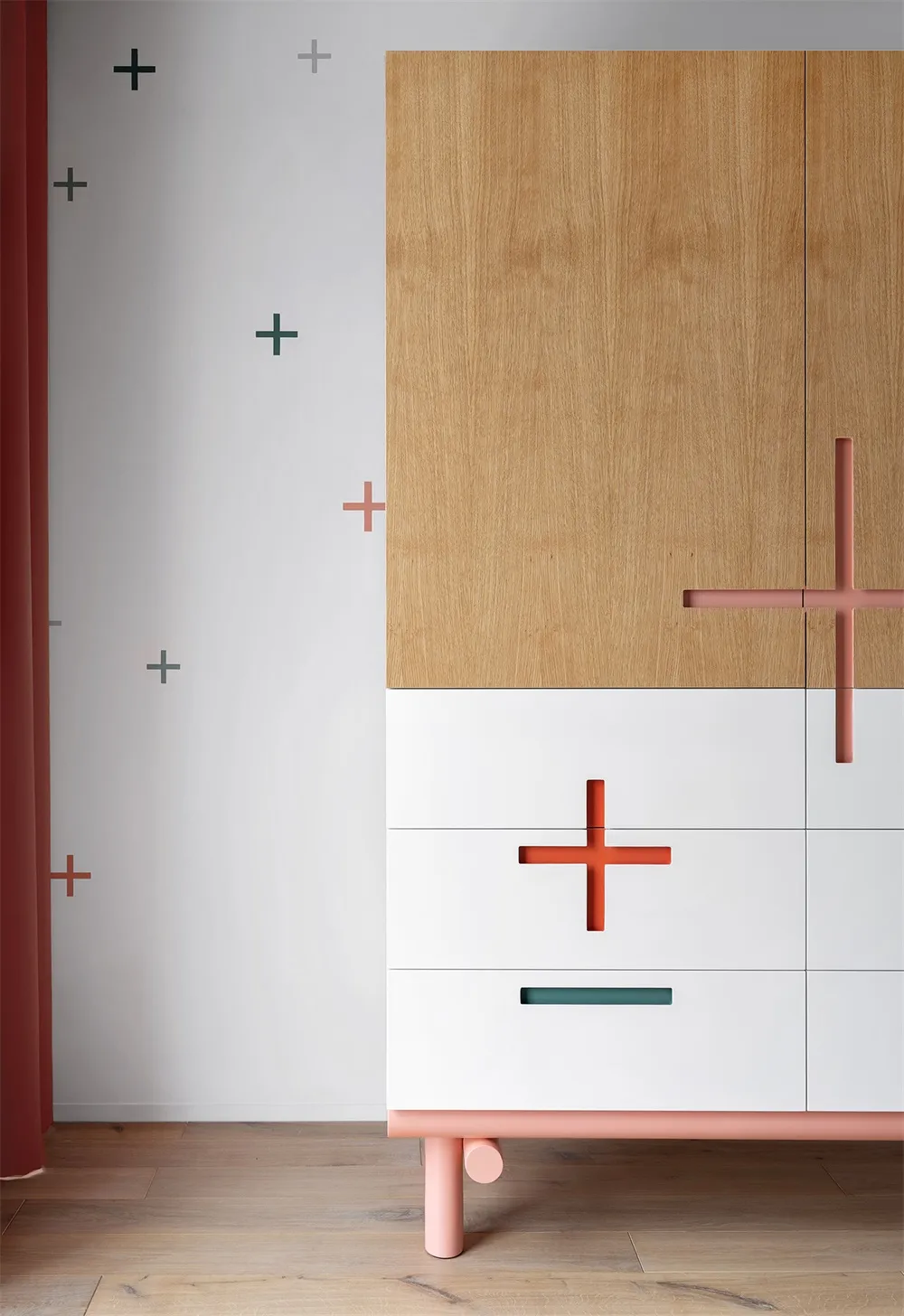 Modern minimalist wardrobe cabinet with wooden and white panels, orange and green crosses, and pink legs.