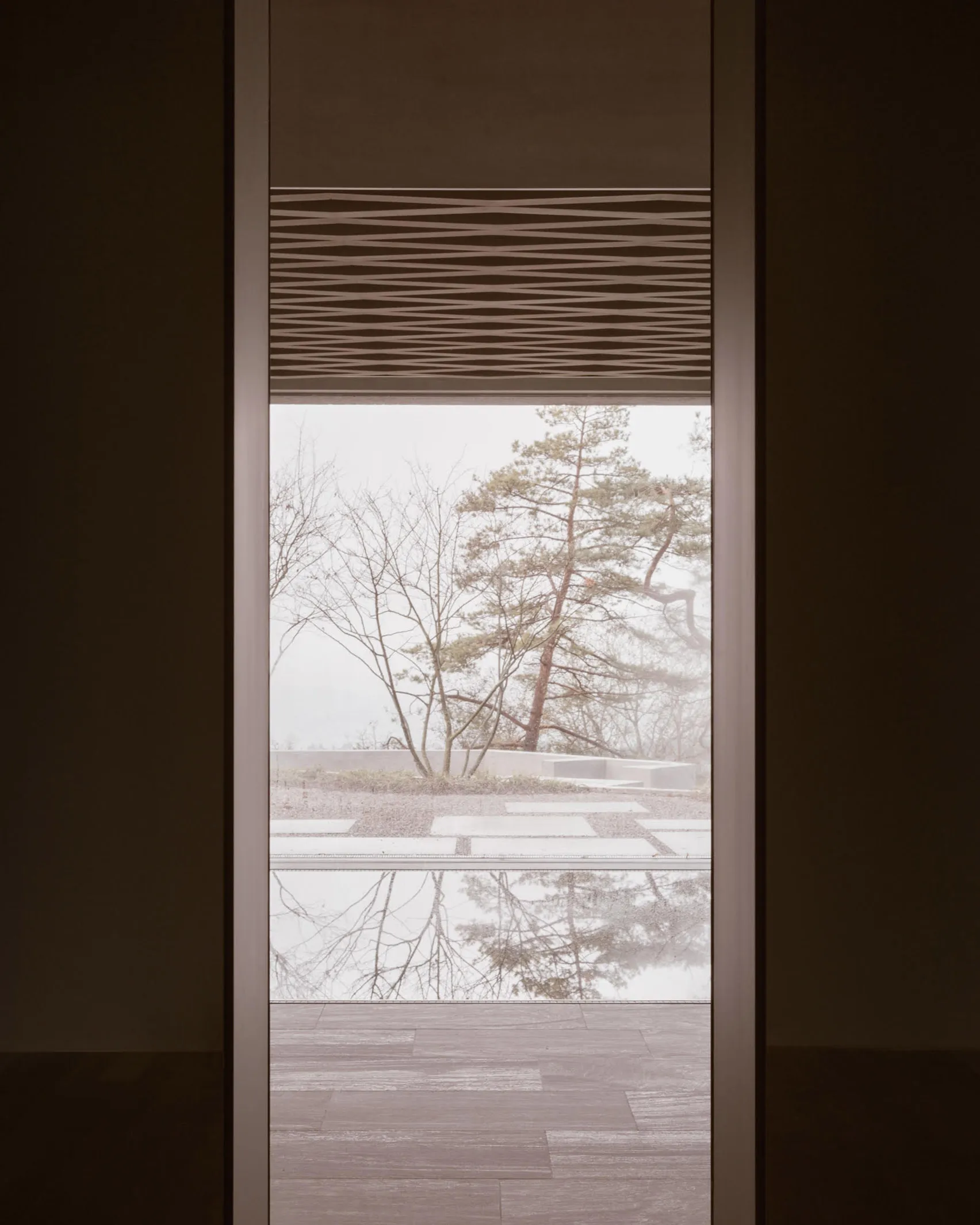 Metal blinds and a finished wooden floor frame a view of nature outside the window.