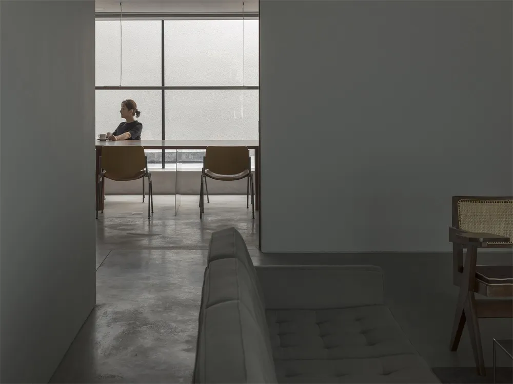 A minimalist modern coffee shop with concrete flooring, a long table, and a woman drinking coffee by a large window.