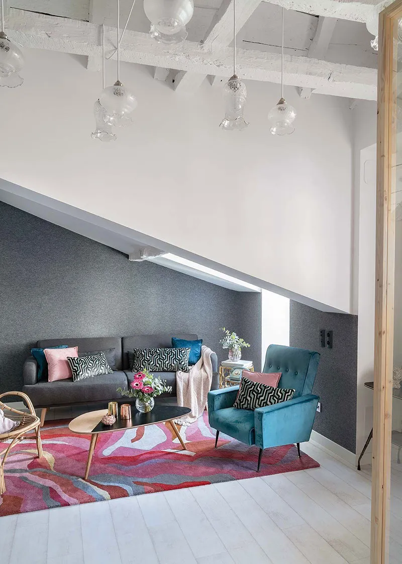 A modern living room featuring a colorful abstract rug, a grey sofa with patterned cushions, a teal armchair, and a black cof