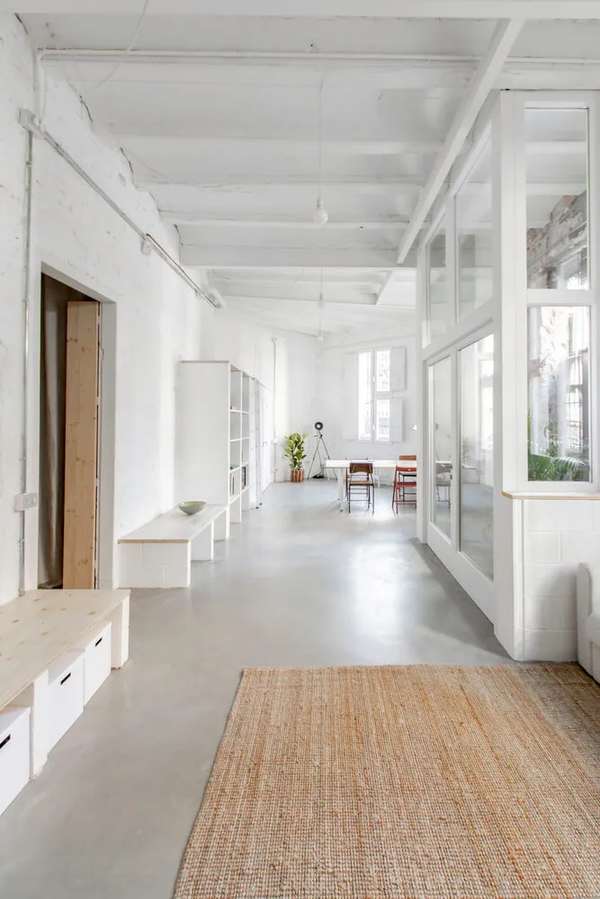 Minimalist workspace with white concrete floors, glass partitions, and simple furniture.