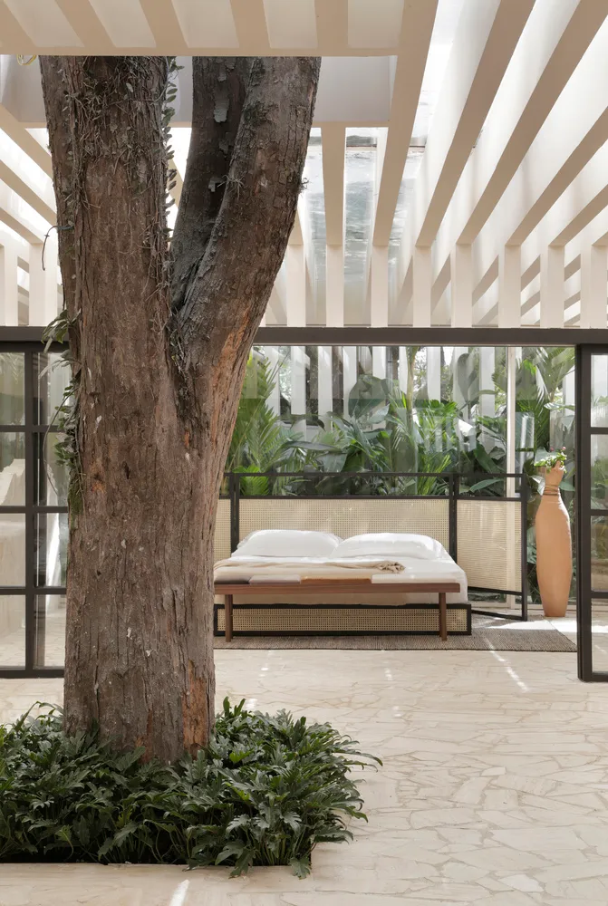 A minimalist bedroom with a white bed and natural wooden elements surrounded by greenery and a tree trunk.