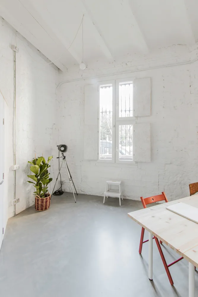 A minimalist office with concrete walls, a white table, and a window with white shutters.