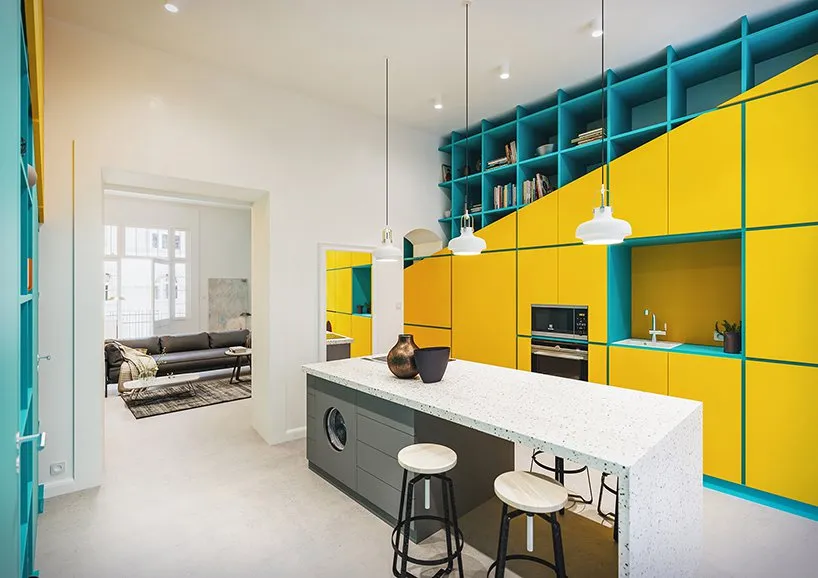 Modern kitchen with yellow and teal cabinets, white countertops, and pendant lights.