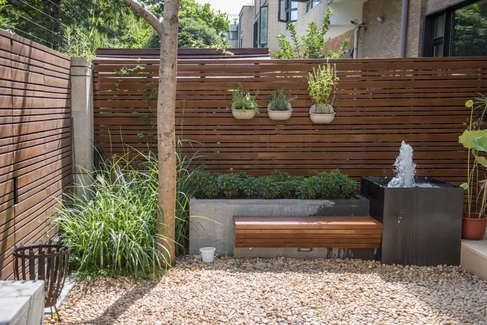 A modern urban garden featuring a wooden fence, concrete seating area, and pebble pathway with a water fountain.