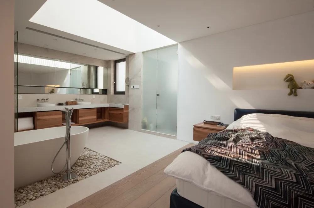 Modern bedroom featuring white walls, a bed with modern bedding, a floating vanity, and a stone-lined bathtub.