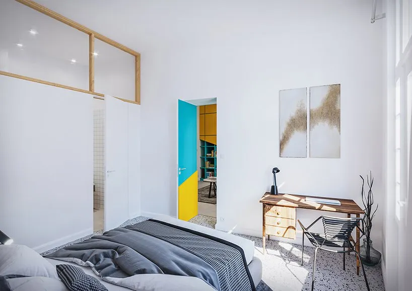 A modern bedroom with white walls, a grey bed, blue and yellow accents, and abstract art on the wall.