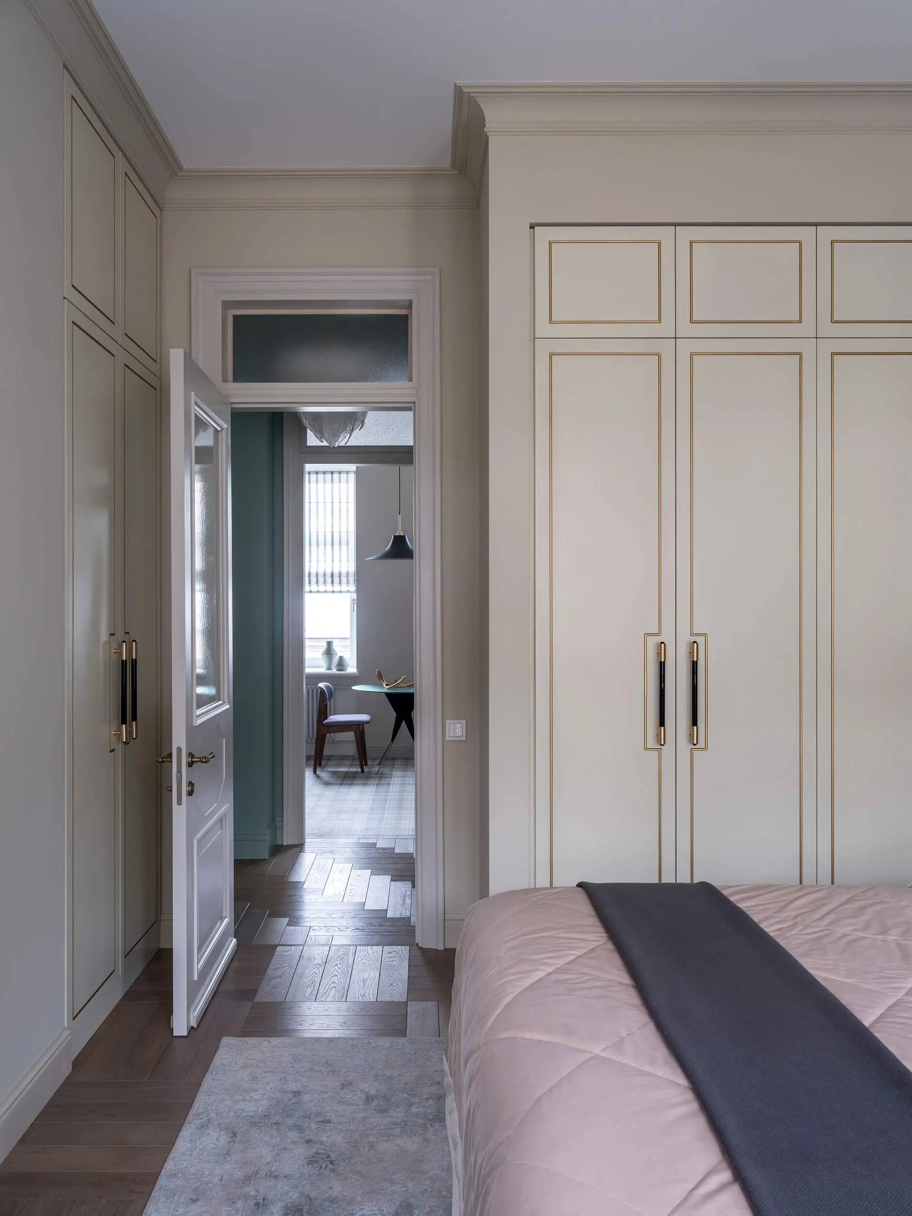 A minimalist bedroom featuring a bed with a pink blanket, a white wardrobe with gold trim, and a wooden floor with a gray rug
