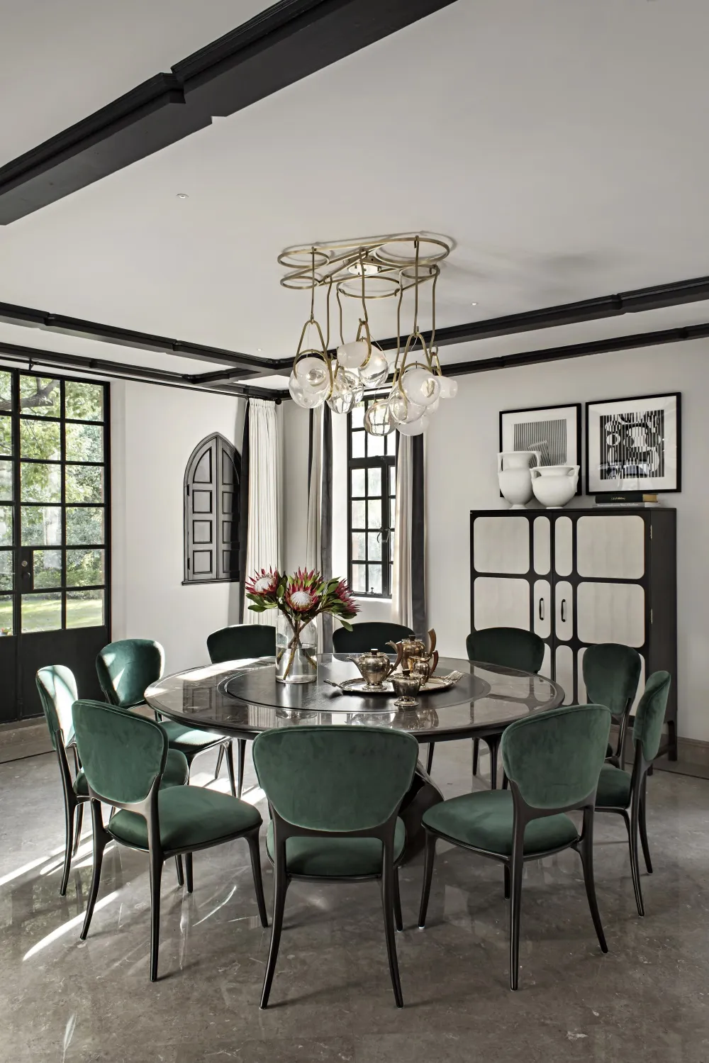 A modern dining room featuring a round table with marble top, surrounded by green velvet chairs with black frames, and a hang
