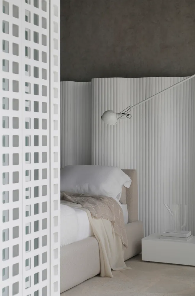 A minimalist bedroom featuring a white bed with a beige throw, a white lamp mounted on the wall, and a white perforated scree