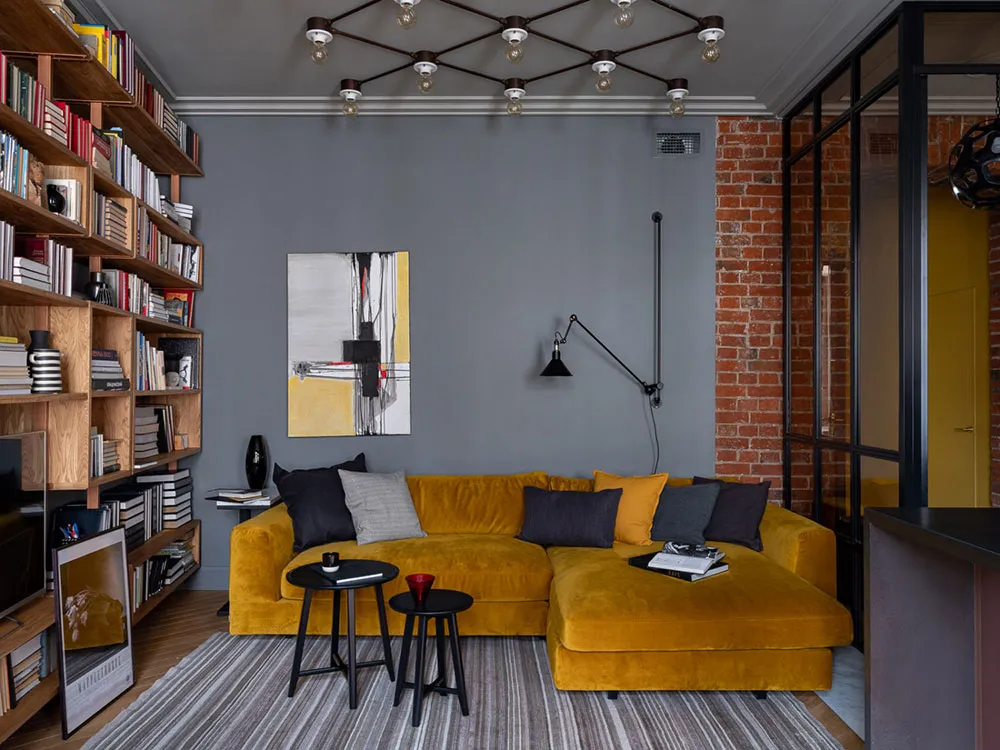 A modern living room featuring a yellow velvet sofa, black stools, and a large bookshelf.