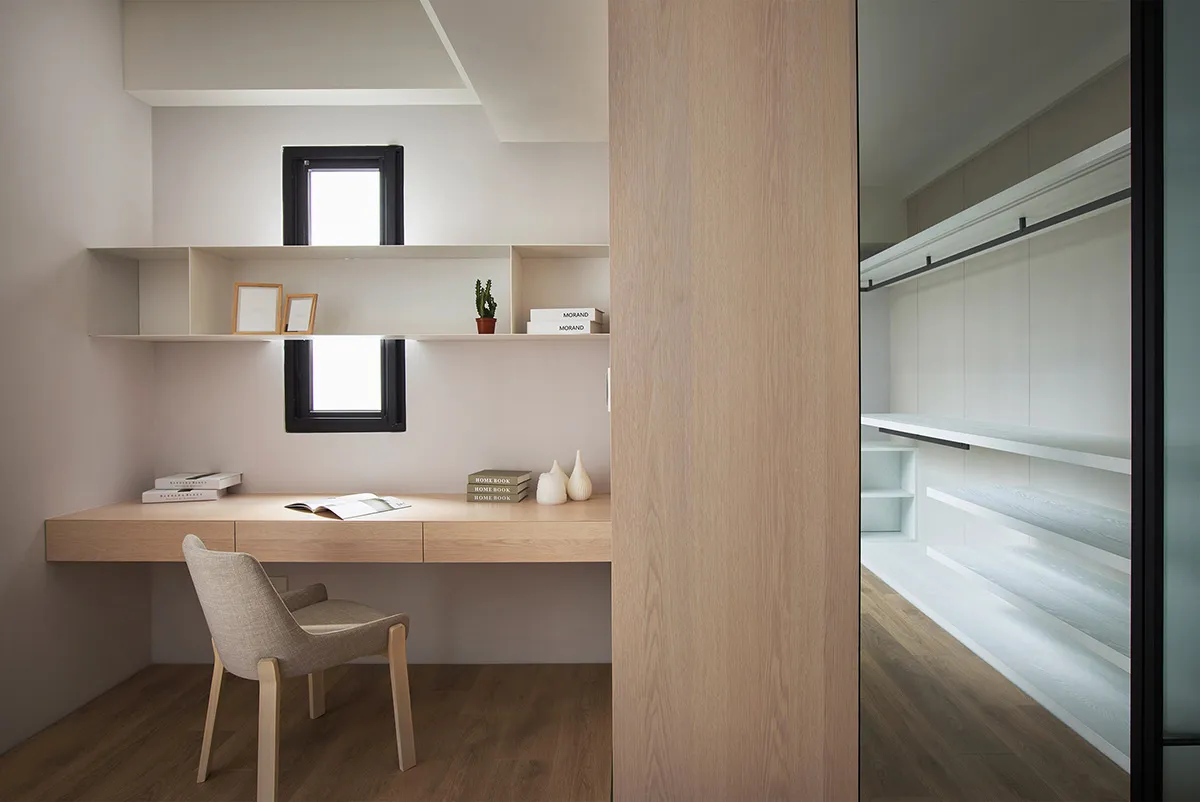 A modern minimalist home office featuring a wooden desk and shelves, with a comfortable chair and a small window.