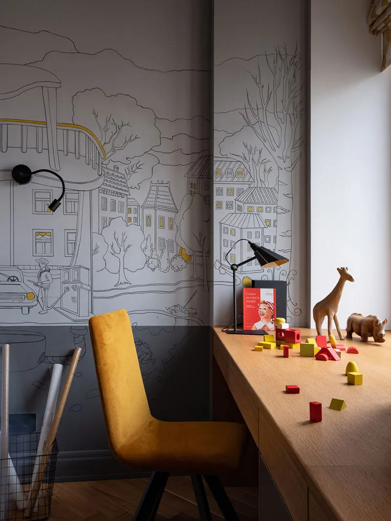 A modern playroom featuring a wooden desk with toys and a mustard yellow chair, against a wall mural depicting a small villag