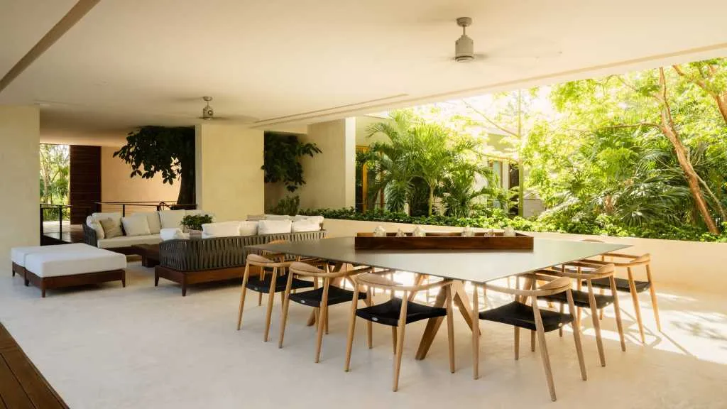 A modern residential interior with white concrete flooring, a long dining table with wooden chairs, and a comfortable seating