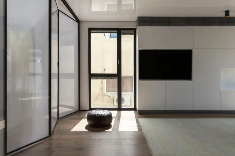 A minimalist living room featuring a large glass door, a cushioned ottoman, and a mounted TV on a white wall.