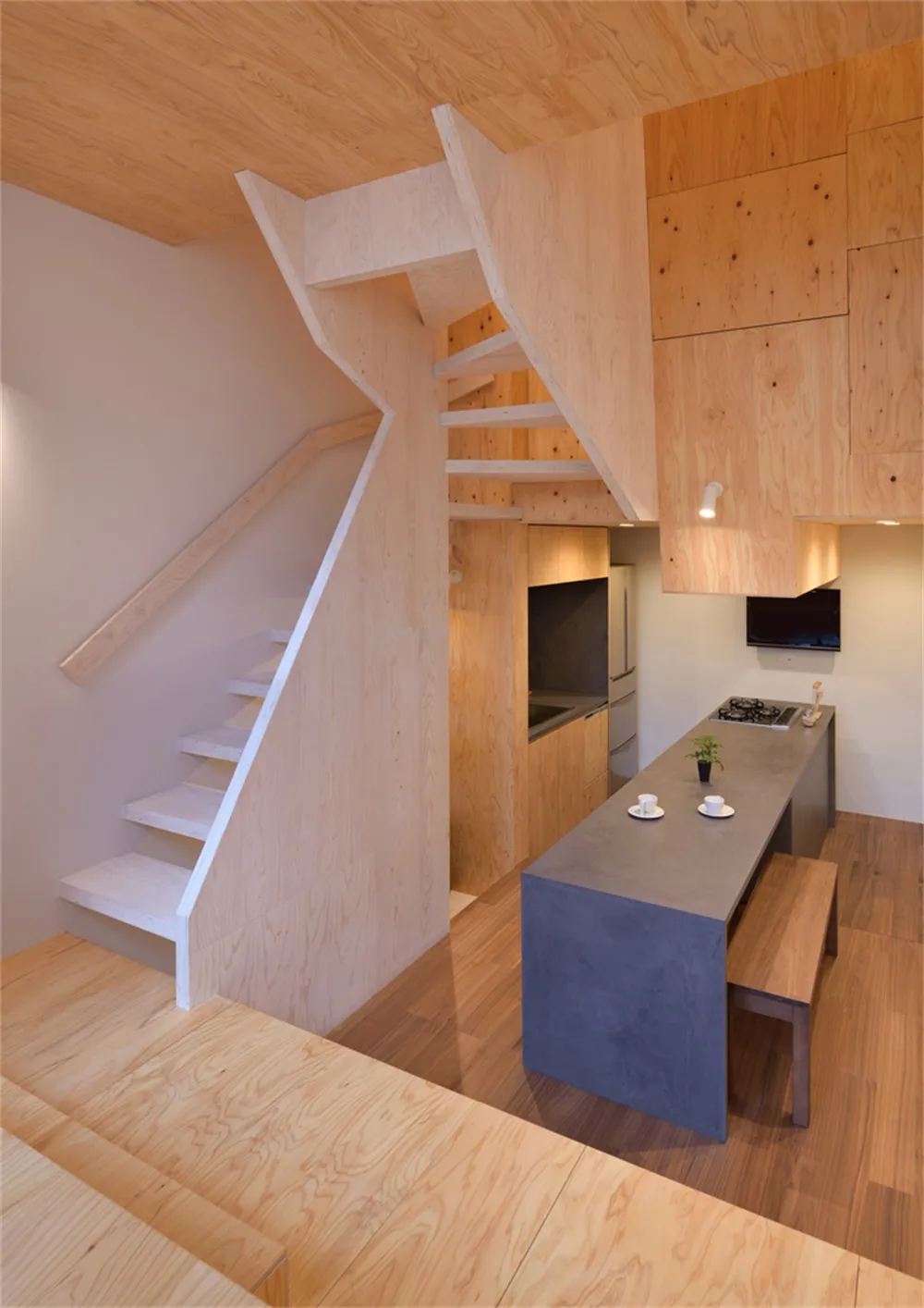 A minimalist residential interior featuring wooden stairs and kitchen area with a concrete island.