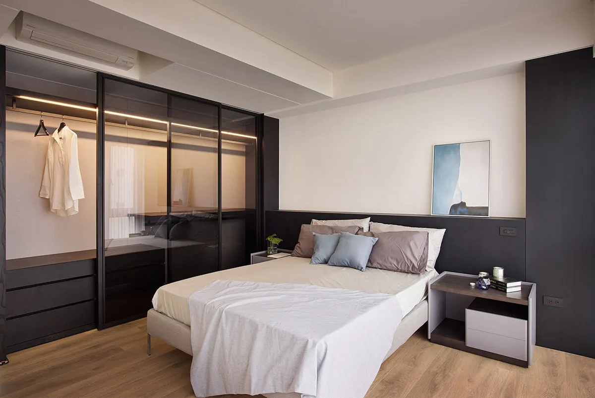 A modern bedroom with minimalist lighting, featuring a bed with white bedding and multiple pillows, a glass wardrobe, and woo
