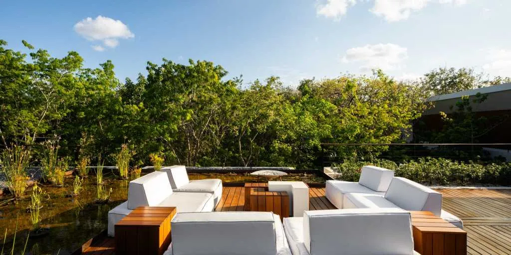 Modern outdoor lounge area with white sofas, wooden tables, and a variety of green plants, set against a backdrop of trees an
