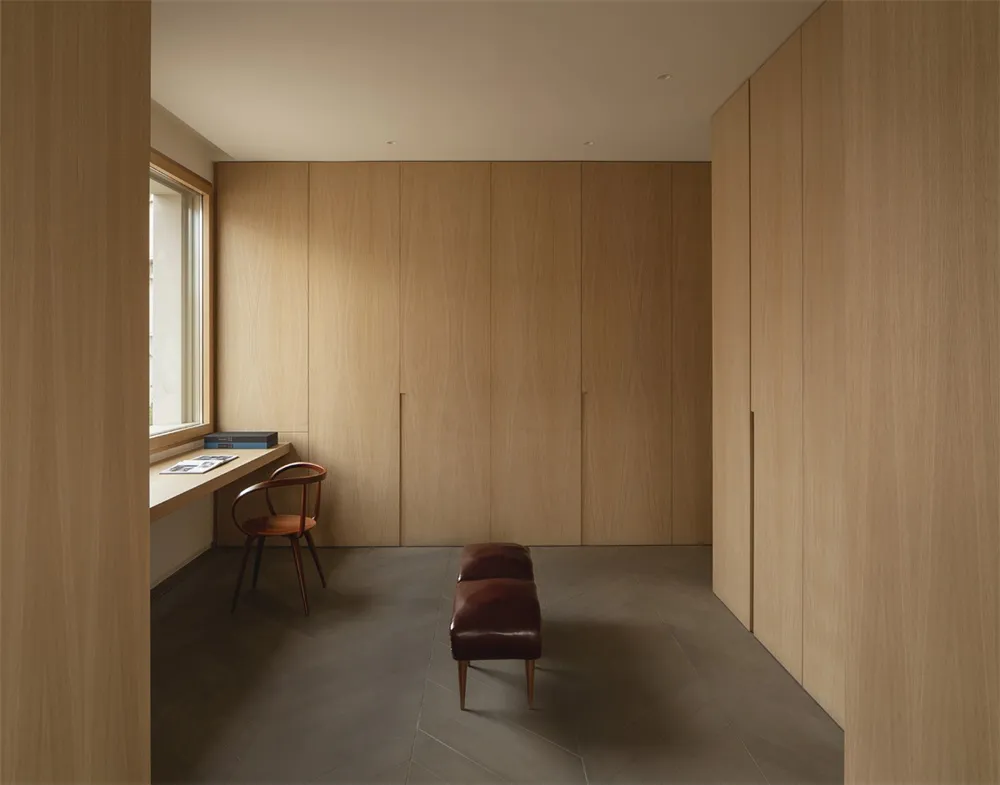 An open, minimalist living area with wooden cabinetry, a bench, and a small desk area by a window.