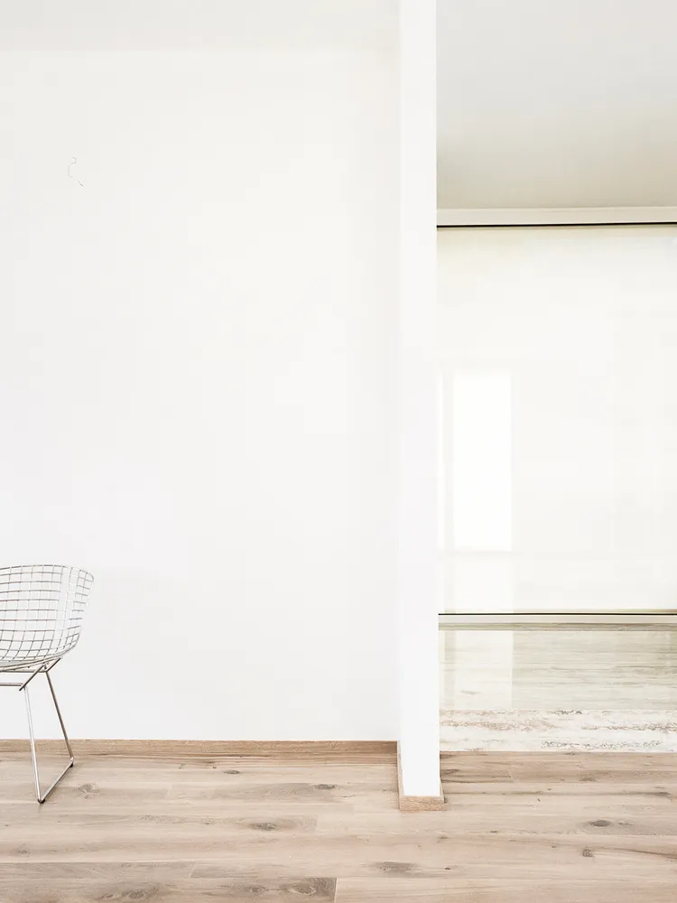 A minimalist room with wooden flooring and a metal wire chair against a white wall.