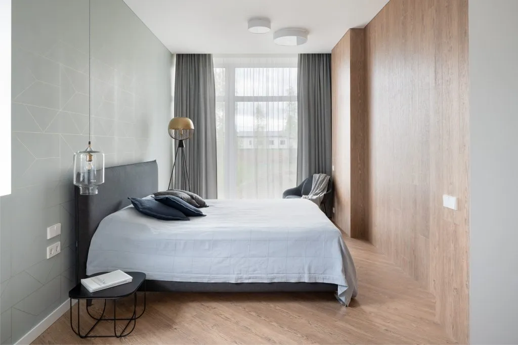 Modern bedroom featuring a grey upholstered bed with navy blue pillows, a bedside table with a lamp, and wooden paneling on o