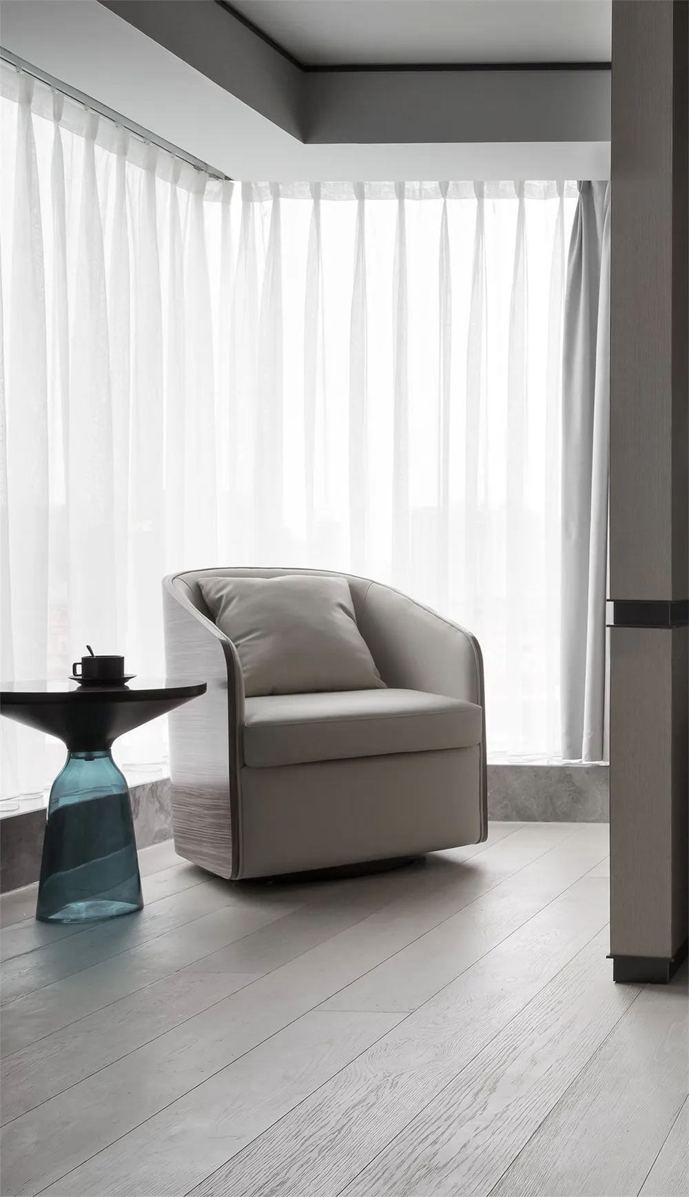 A modern residential space featuring a beige upholstered chair with a glass table and sheer curtains by the window.