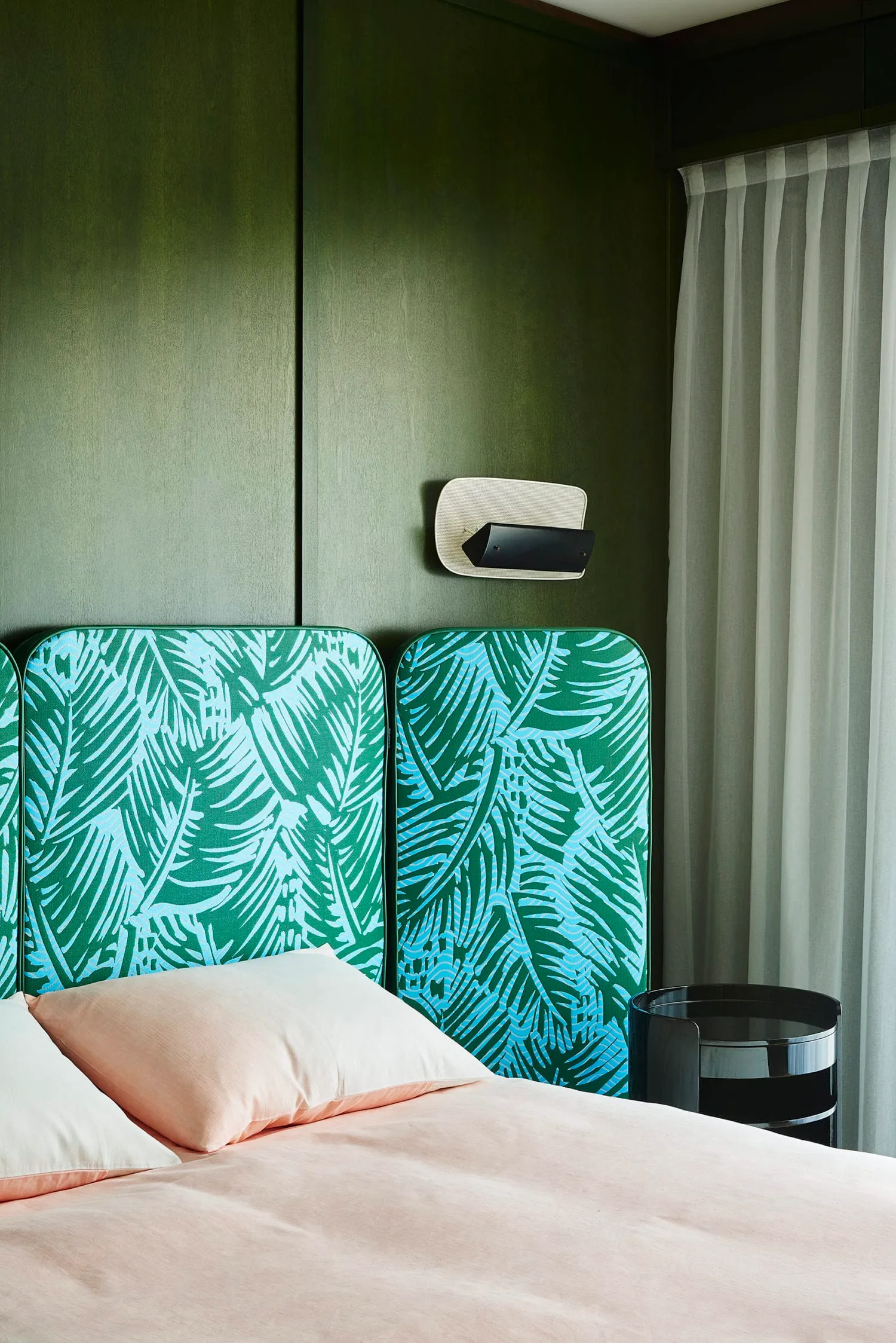 A modern bedroom featuring a bed with pink bedding, a green palm leaf print headboard, and a window with sheer curtains.