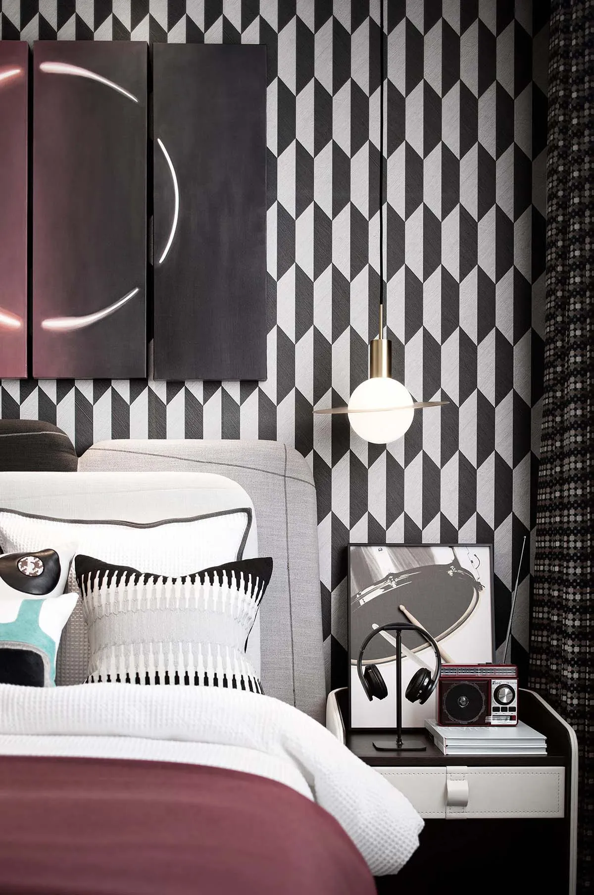 A modern bedroom featuring a geometric patterned backdrop, a bed with a white headboard and burgundy accents, and a hanging l