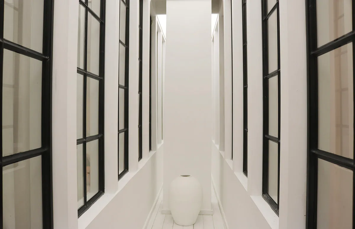 A minimalist hallway with white walls and windows, featuring a single white vase.