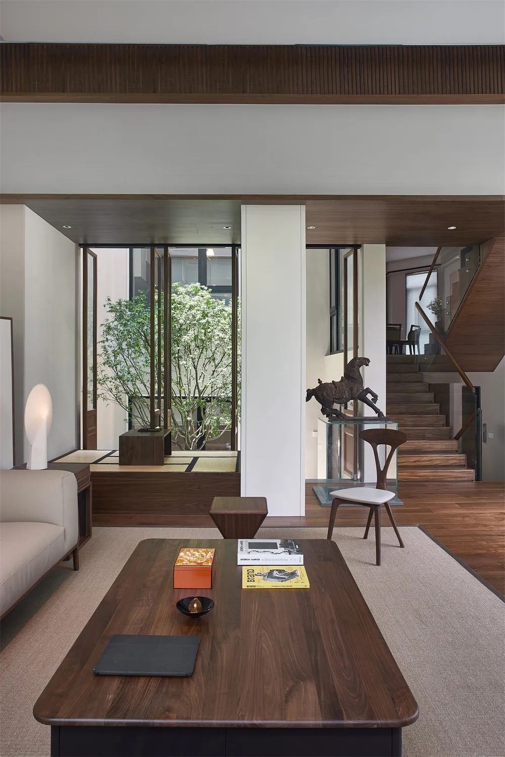 A modern living room with wooden furniture, featuring a large window, a coffee table with books and a bowl, and a wooden floo