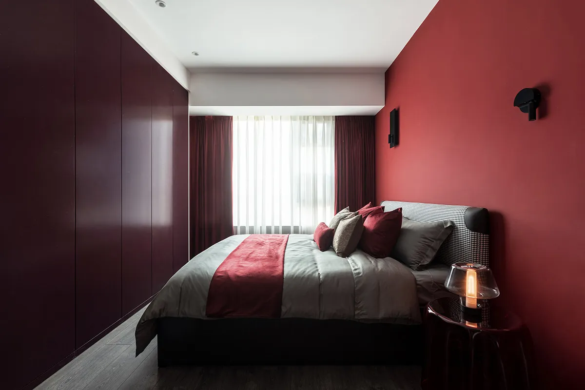 A modern bedroom with a red accent wall, bed with mixed textiles, and a sleek, minimalist design.