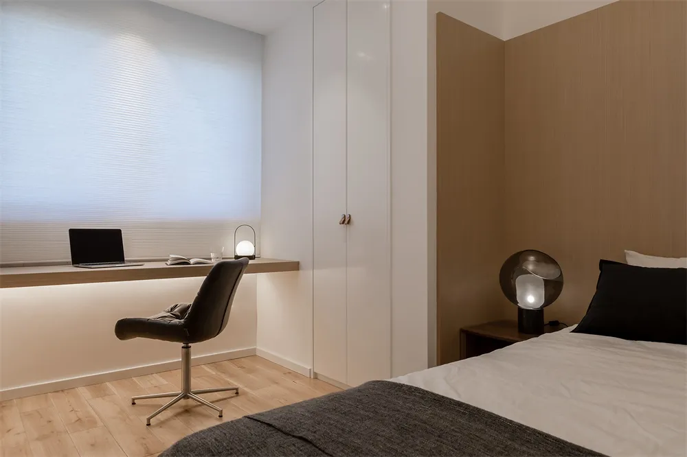 A minimalist bedroom with a wooden floor, a black office chair with a round base, a desk holding a laptop and a lamp, a bed w