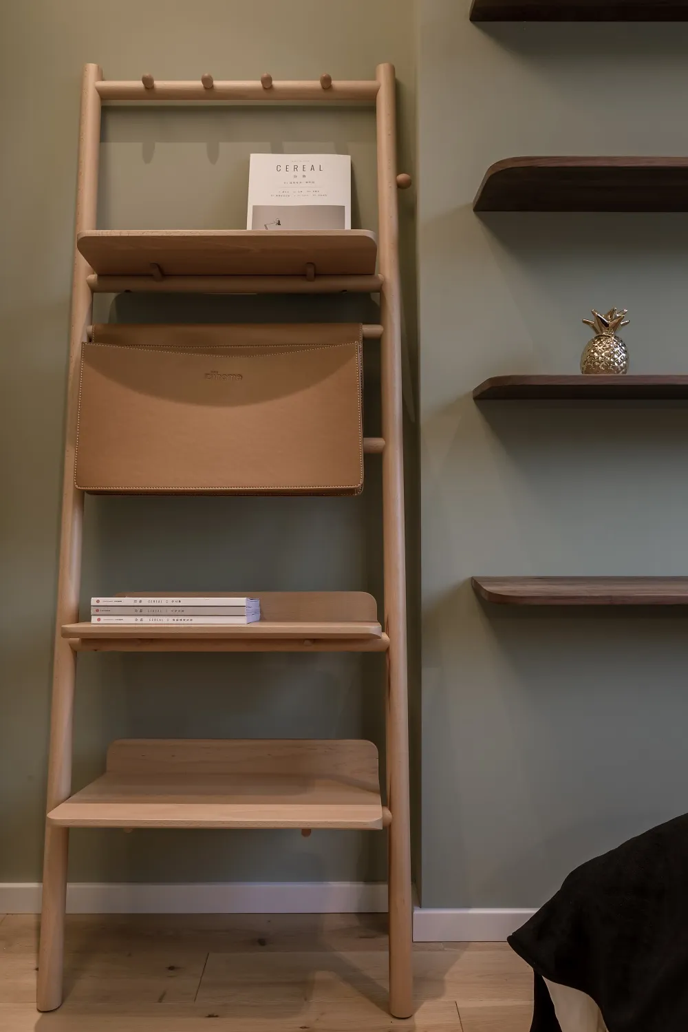A wooden bookshelf with shelves and a bag attached to it, featuring a book and a golden pineapple ornament.