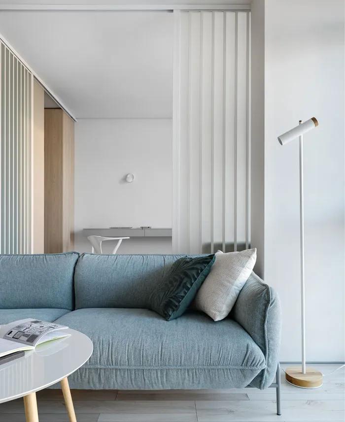 A minimalist living room featuring a blue fabric sofa, white wooden floor lamp, and white walls.