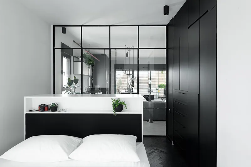 A minimalist bedroom features a sleek, black-and-white color palette, with a glass partition, wooden flooring, and metal fixt